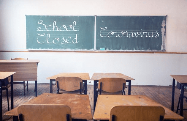 This image from a primary schoolroom features an empty teacher's desk and several student desks with chairs, all setting empty. At the front of the classroom there are two chalkboards, one of which reads "School Closed" and the other of which reads, "Coronavirus."